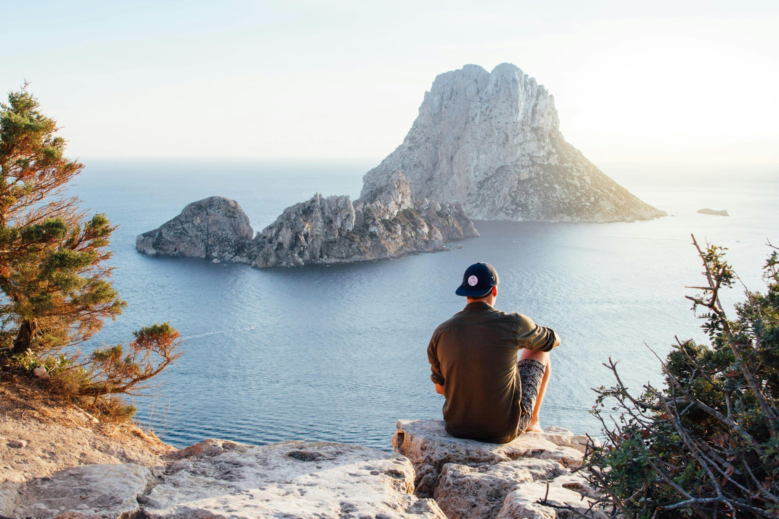 Man enjoys a scenic view of Es Vedrà at sunset from a cliff in San Juan Bautista, providing a perfect summer escape.